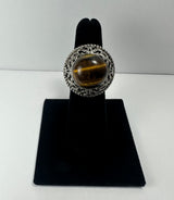 Silver ring featuring a round tiger's eye gemstone with a golden-brown chatoyant band, set in an ornate filigree bezel on a black ring display.