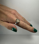 Hand with dark green nail polish wearing a hexagonal quartz ring with a silver band, shown against a plain light gray background.