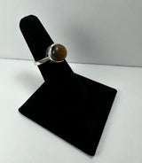 Silver ring with a round, polished brown Tiger's Eye stone set in a simple bezel, displayed on a black velvet ring holder against a white background.