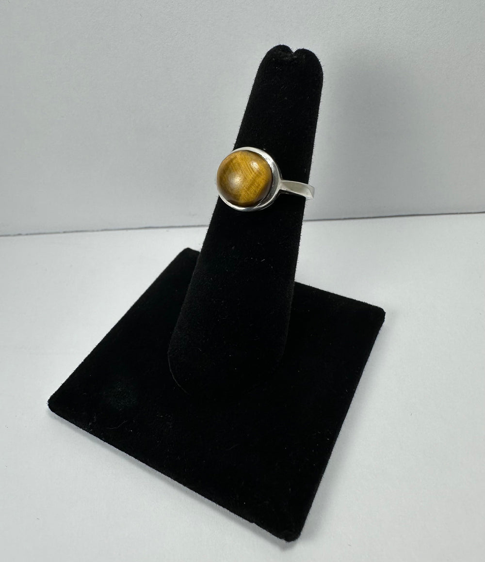 Silver ring with a polished round tiger's eye stone, displaying golden-brown chatoyant stripes, mounted on a black velvet finger display stand.
