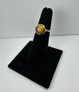 Silver ring with a polished round tiger's eye stone, displaying golden-brown chatoyant stripes, mounted on a black velvet finger display stand.