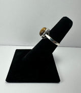 Silver ring with a polished round tiger's eye stone set in a simple bezel, displayed on a black velvet ring holder against a plain background.
