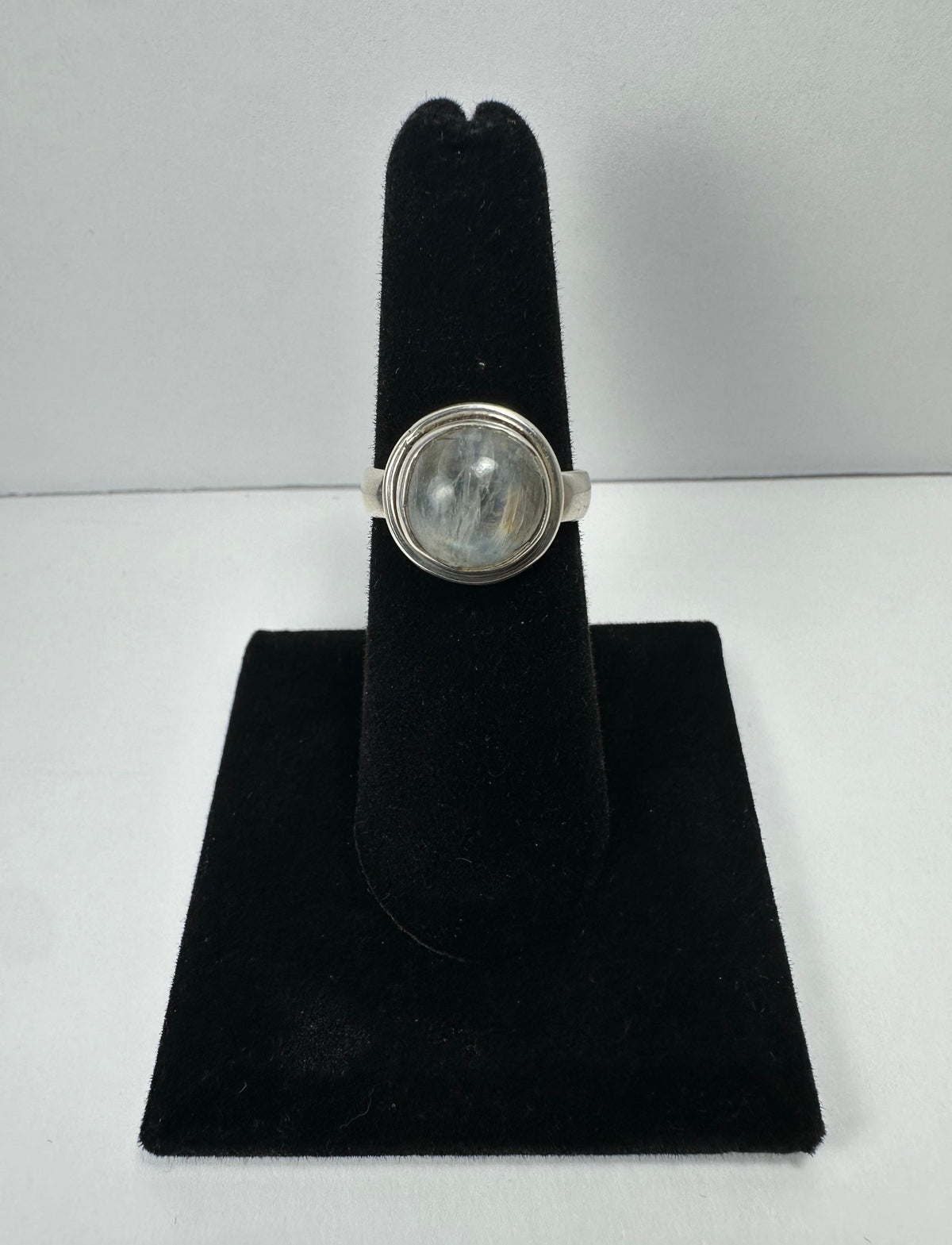 Round moonstone cabochon set in a simple silver bezel ring displayed on a black velvet ring holder with a plain white background.