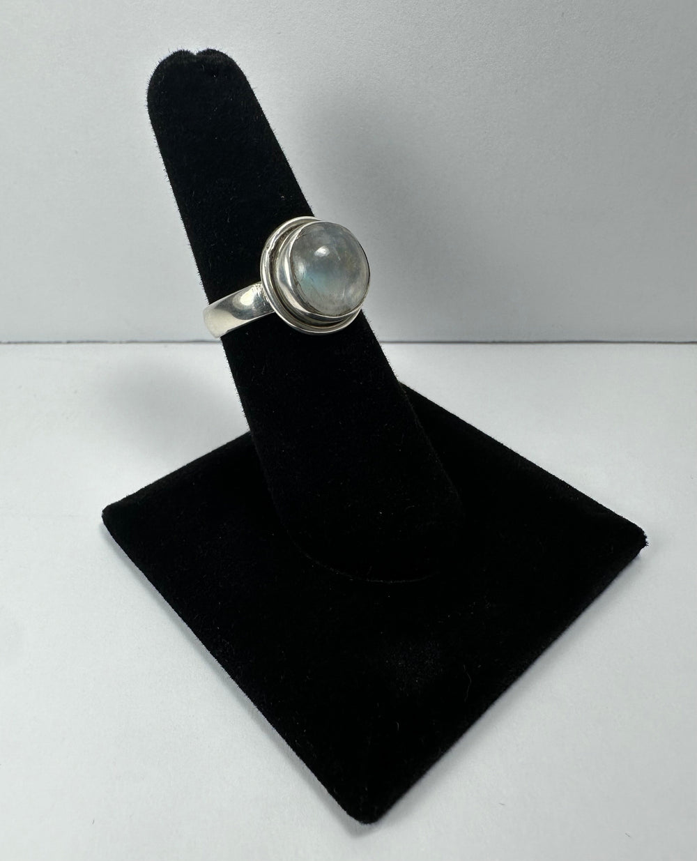 Silver ring with a round, translucent moonstone cabochon set in a simple bezel, displayed on a black velvet ring holder against a plain gray background.