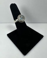 Silver ring with a round, translucent moonstone cabochon set in a simple bezel, displayed on a black velvet ring holder against a plain gray background.