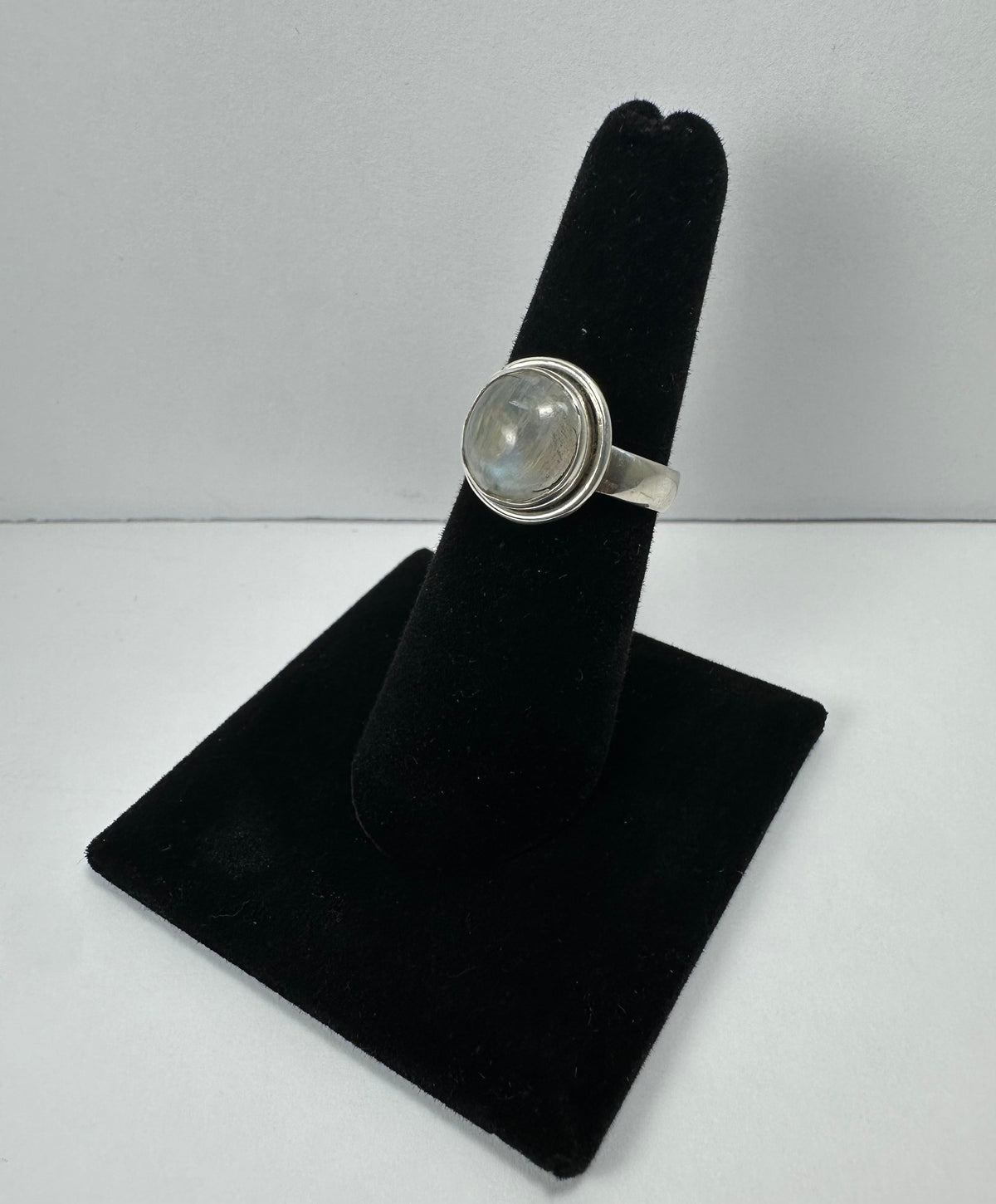 Round moonstone set in a silver bezel ring, displayed on a black velvet finger stand against a plain background.