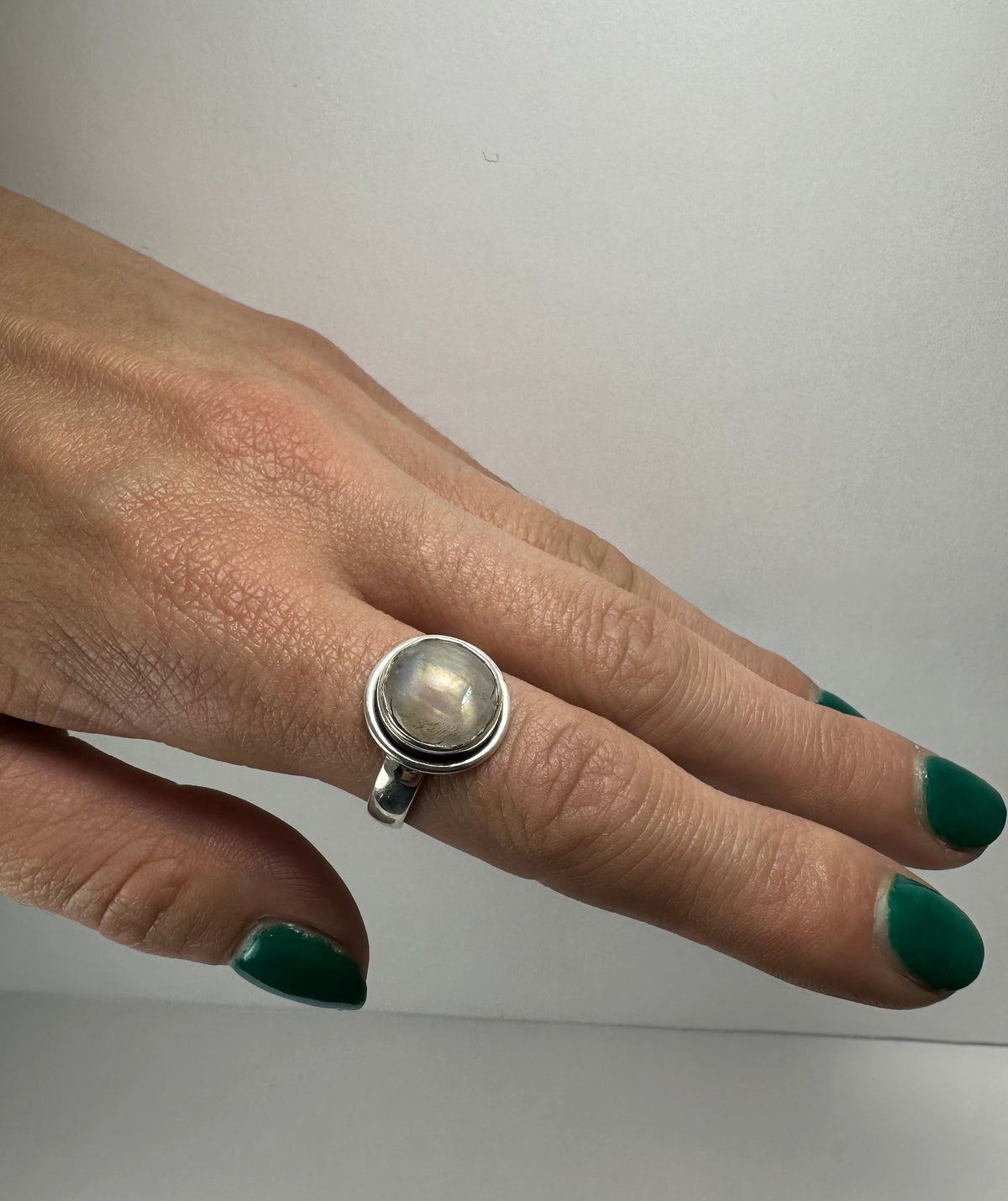 Silver ring with a round, translucent moonstone set in a bezel, worn on a hand with dark green painted nails against a plain background.
