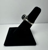 Silver ring with a round, polished black onyx stone set in a simple bezel, displayed on a black velvet finger stand against a light background.