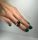 Hand with green nail polish wearing a silver ring featuring a round black onyx stone, shown against a plain light gray background.