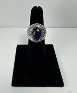 Silver ring with a rounded deep purple amethyst stone center and textured geometric pattern surrounding the stone, displayed vertically on black velvet stand.