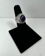 Silver ring with a large oval purple amethyst cabochon set in the center, featuring a textured geometric pattern on the band, displayed on a black velvet ring stand.