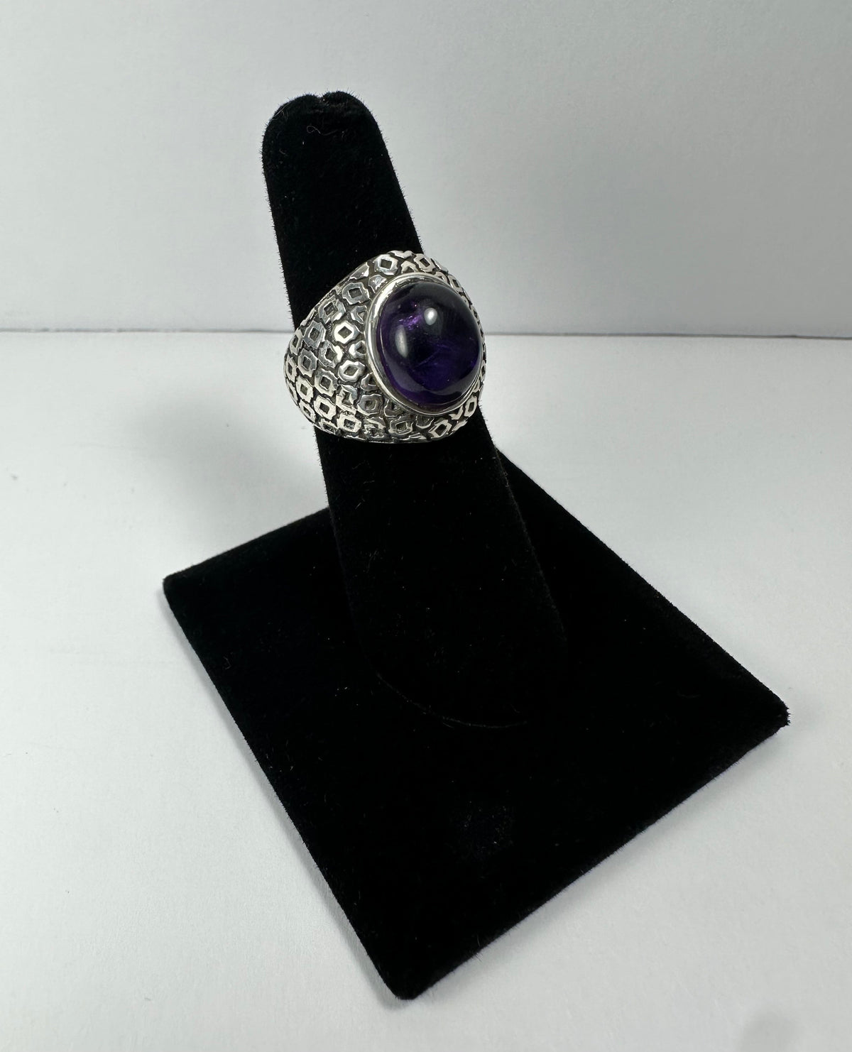 Silver ring with a large oval purple amethyst cabochon set in the center, featuring a textured geometric pattern on the band, displayed on a black velvet ring stand.