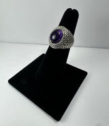 Silver ring with an oval cabochon amethyst center stone and a textured, geometric patterned band displayed on black velvet stand.