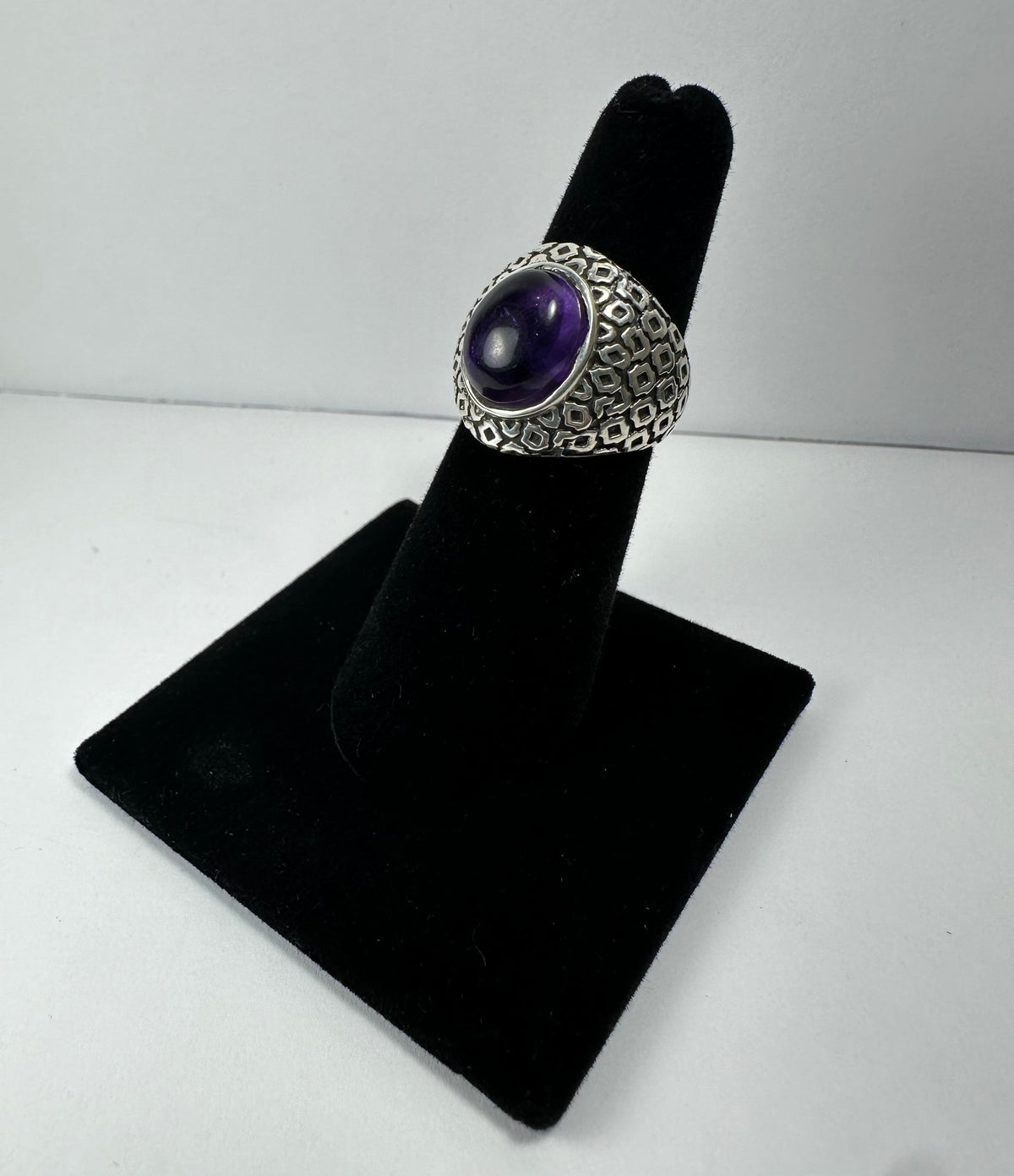 Silver ring with an oval cabochon amethyst center stone and a textured, geometric patterned band displayed on black velvet stand.