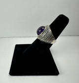 Silver ring with a rounded purple amethyst stone set in a wide band featuring a geometric hexagonal pattern, displayed on a black velvet ring holder.