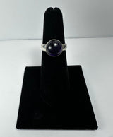 Silver ring with a round, polished deep purple amethyst stone set in a bezel, displayed on a black velvet finger stand against a white background.