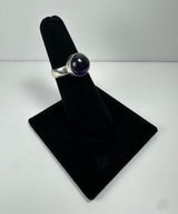Silver ring with a smooth, round deep purple amethyst stone set in a simple bezel, displayed upright on a black velvet ring stand against a light background.