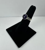 Silver ring with a polished round amethyst stone mounted on a black velvet ring display stand against a plain white background.