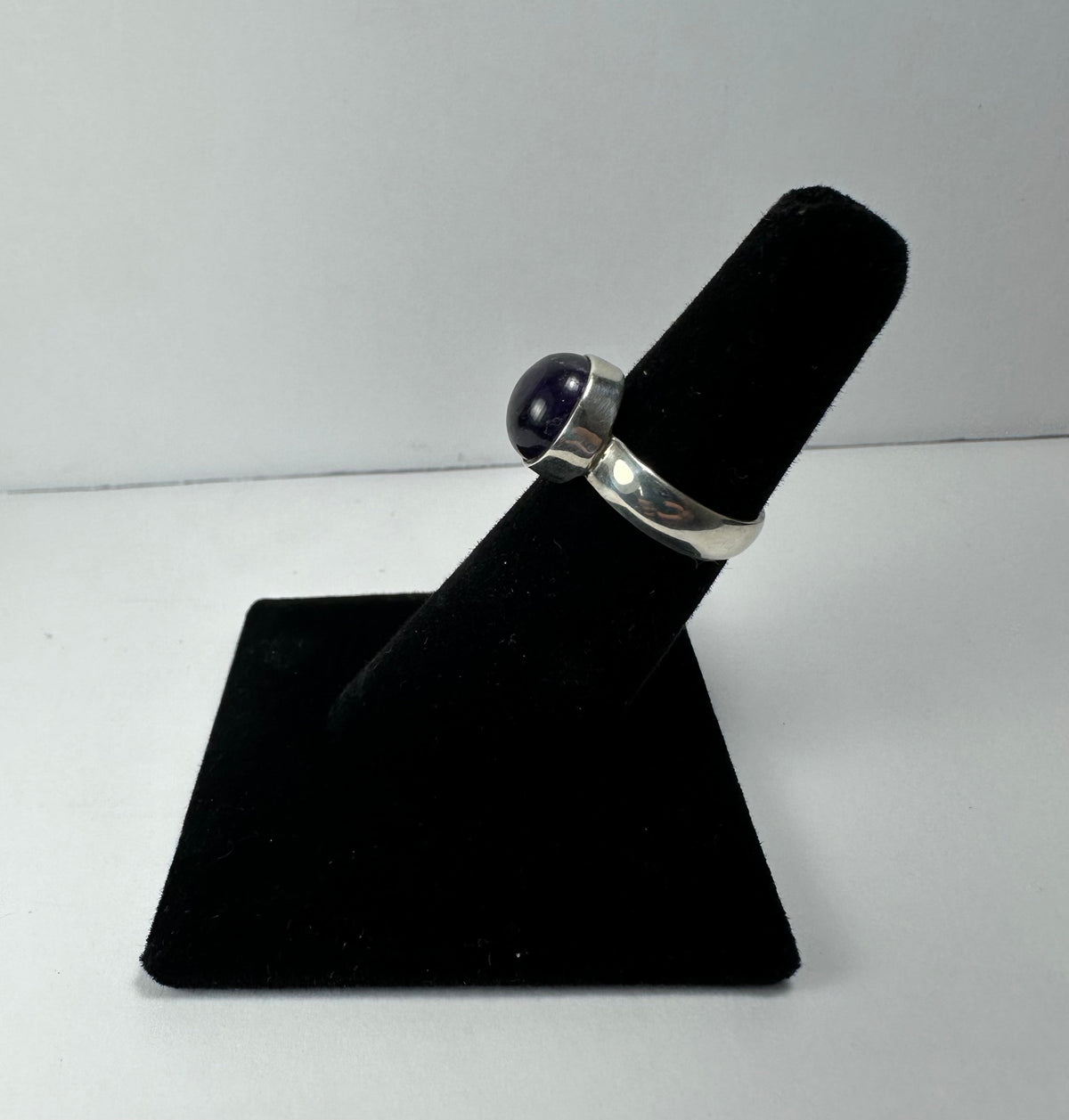 Silver ring with a round, polished amethyst gemstone set in a bezel, displayed on a black velvet ring holder against a white background.