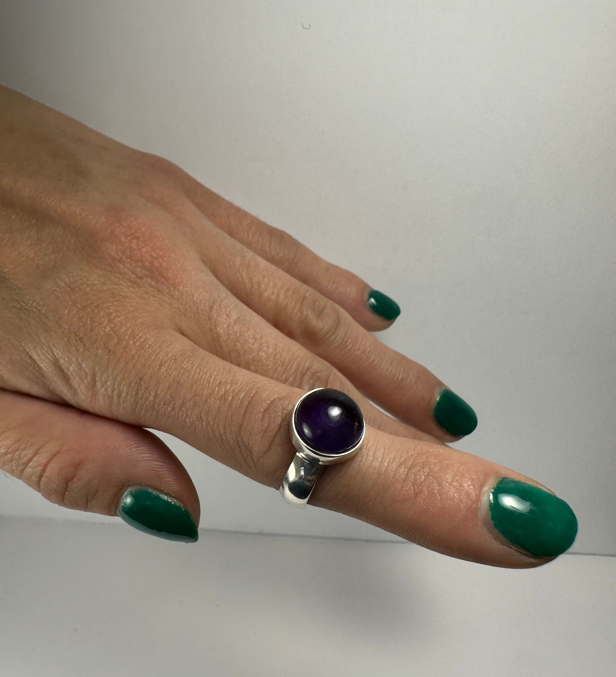 Close-up of a hand with green nail polish wearing a silver ring featuring a round, polished deep purple amethyst stone on the middle finger against a plain light background.