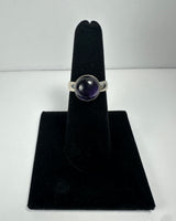Silver ring with a round, polished deep purple amethyst stone set in a simple bezel, displayed on a black velvet finger stand against a plain white background.