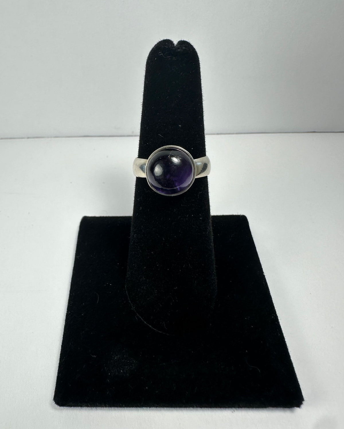 Silver ring with a round, polished deep purple amethyst stone set in a simple bezel, displayed on a black velvet finger stand against a plain white background.