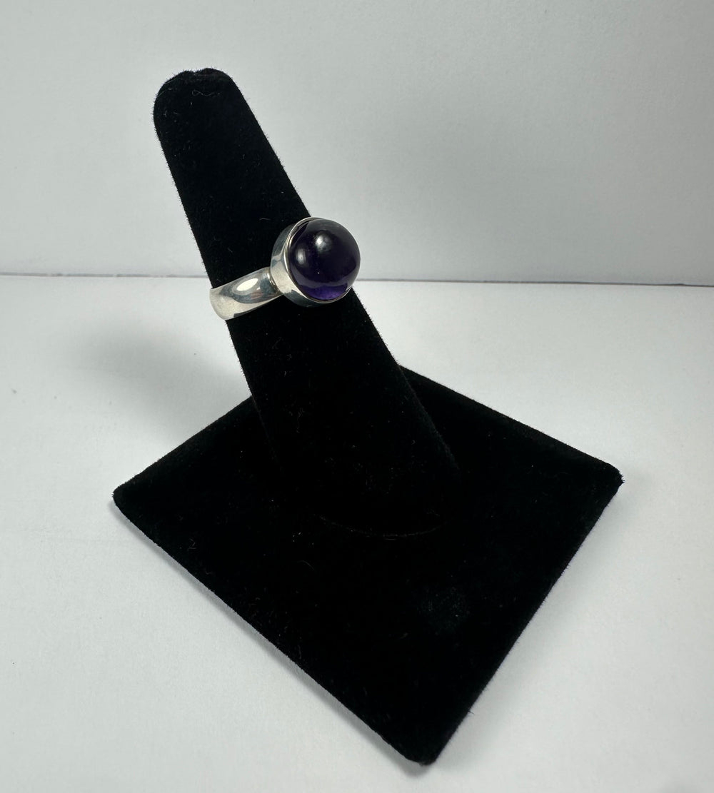 Silver ring with a round, deep purple amethyst stone set in a bezel, displayed on a black velvet ring stand against a neutral background.