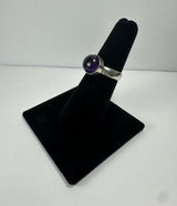 Silver ring with a polished round deep purple amethyst stone set in a simple bezel mount, displayed on a black velvet finger stand against a plain white background.