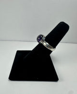 Silver ring with a polished deep purple amethyst cabochon set in a simple bezel setting, displayed on a black velvet ring holder against a white background.