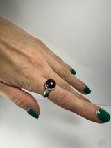 Silver ring with a round, deep purple amethyst stone worn on a hand with green-painted nails against a plain white background.