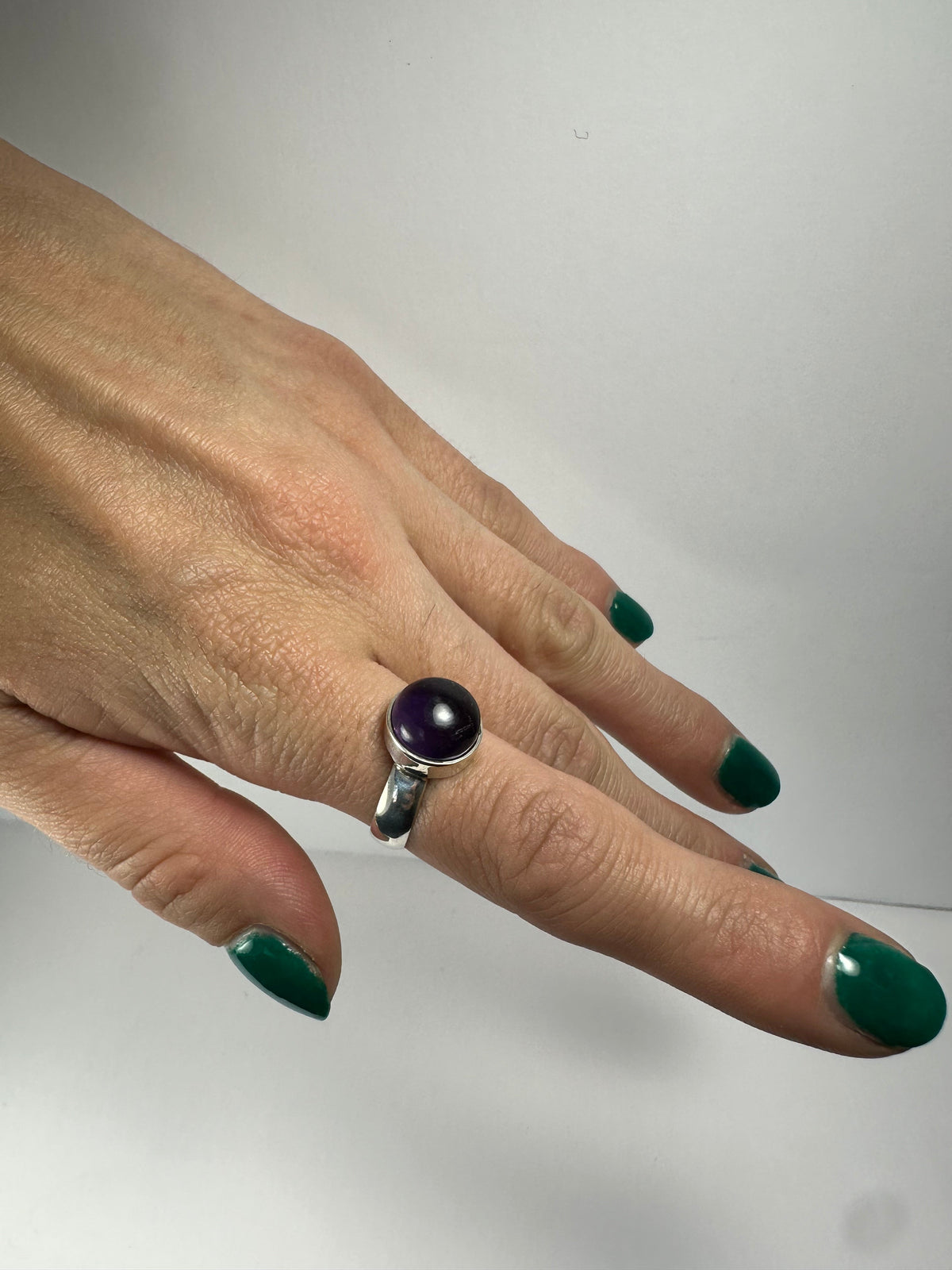 Silver ring with a round, deep purple amethyst stone worn on a hand with green-painted nails against a plain white background.