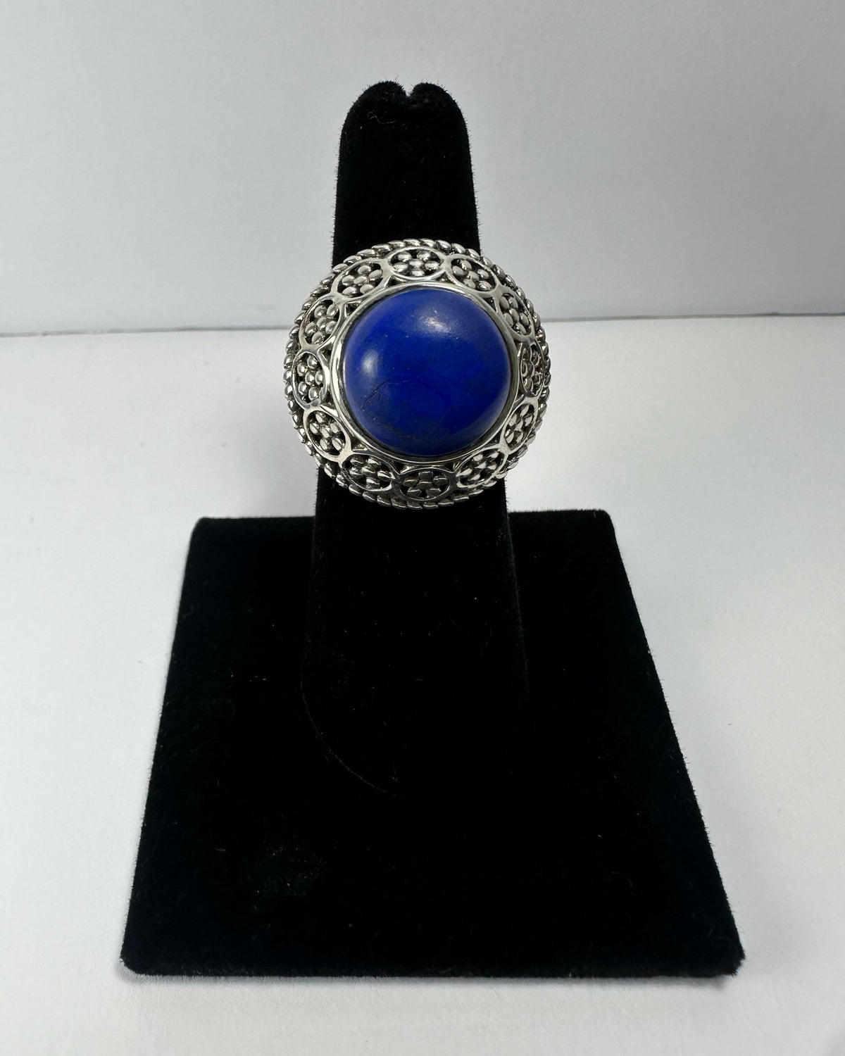 Silver ring with a large round deep blue lapis lazuli stone surrounded by intricate filigree detailing, displayed on a black velvet finger stand.