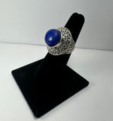 Silver ring with a large round deep blue lapis lazuli stone set in an ornate filigree band, displayed upright on a black velvet ring holder.