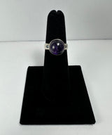 Silver ring featuring a polished round amethyst gemstone set in a smooth bezel on a black velvet display stand against a plain white background.