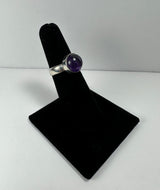 Silver ring with a round purple amethyst stone set in a bezel setting, displayed on a black velvet ring stand against a white background.