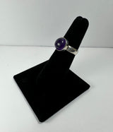 Silver ring with a round purple amethyst stone set in a bezel on a black velvet ring display stand against a white background.