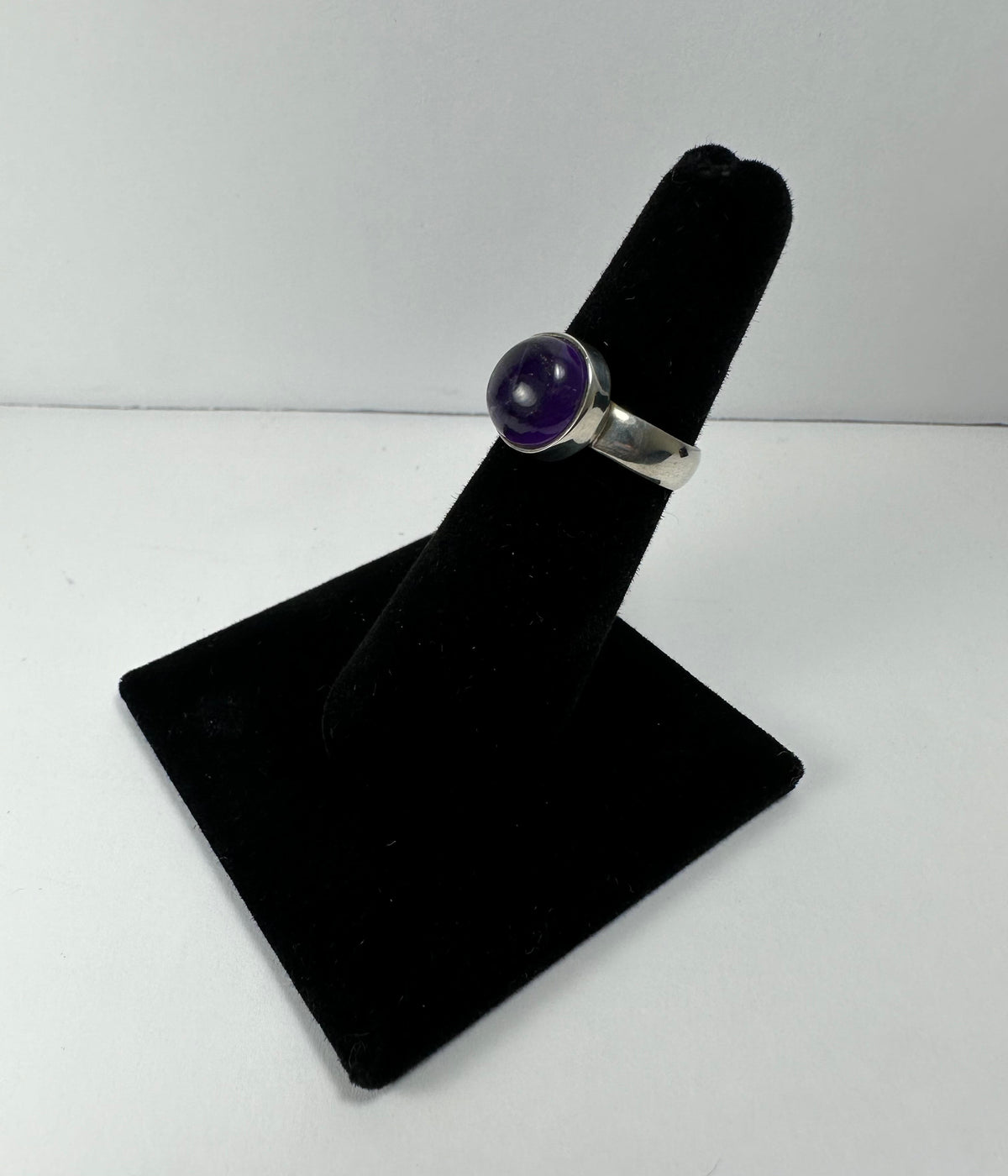 Silver ring with a round purple amethyst stone set in a bezel on a black velvet ring display stand against a white background.