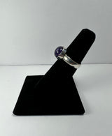 Silver ring with a round, polished amethyst stone mounted on a simple bezel setting, displayed on a black velvet ring stand against a white background