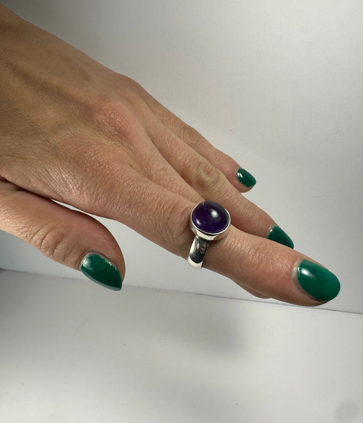 Hand with green nail polish wearing a silver ring featuring a round purple amethyst stone set in a bezel setting on the middle finger.