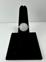 Round moonstone ring with a translucent pale blue gemstone set in a simple silver band displayed on a black velvet stand against a white background.