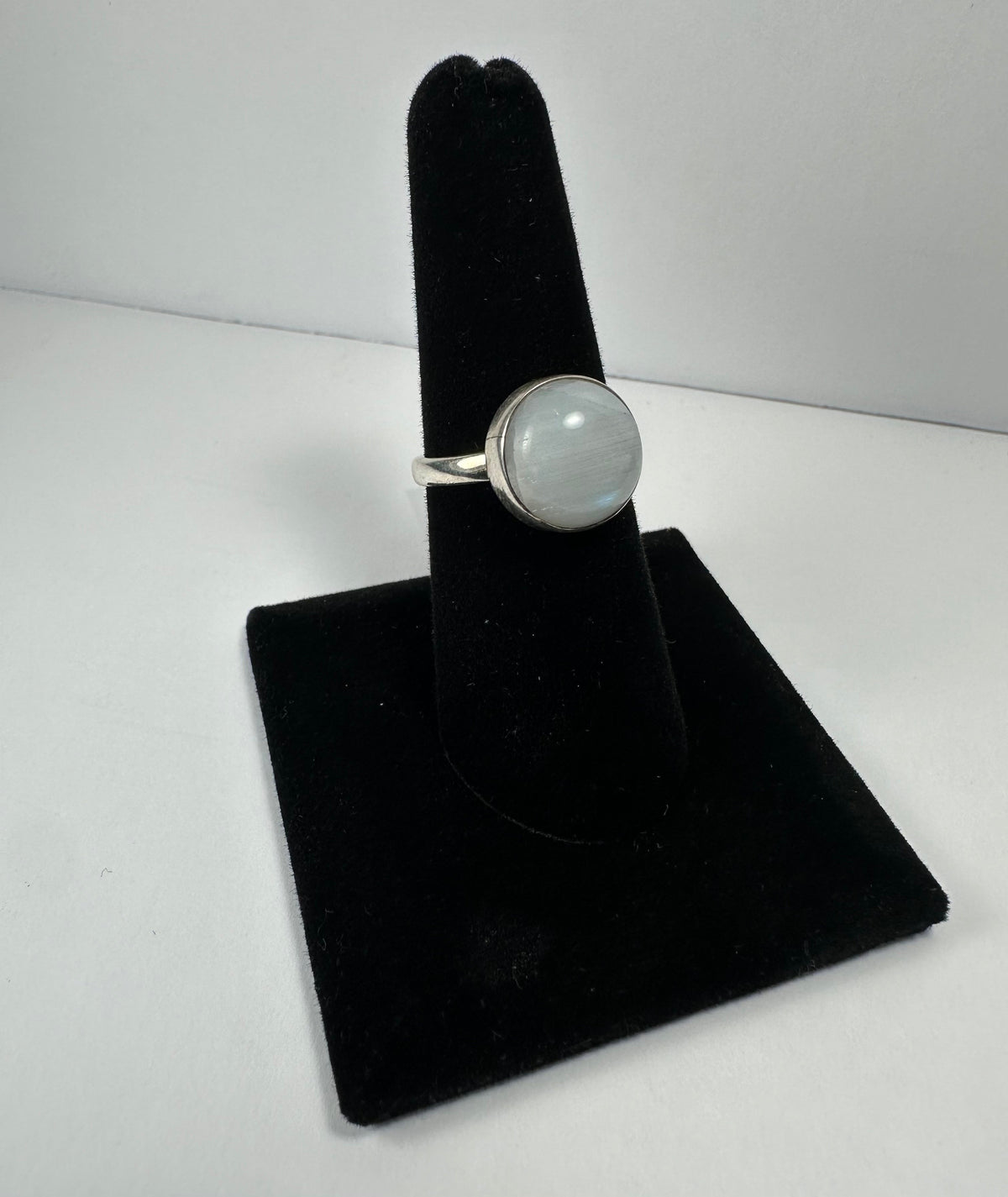 Round moonstone set in a silver bezel on a simple silver band, displayed on a black velvet ring holder against a white background.