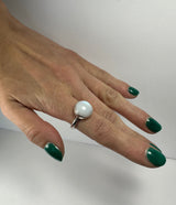 Hand with green nail polish wearing a silver ring featuring a round, smooth, white moonstone on the middle finger against a plain light background.