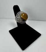 Silver ring with intricate floral and circular patterns featuring a polished brown tiger's eye stone, displayed on a black velvet ring stand against a white background.