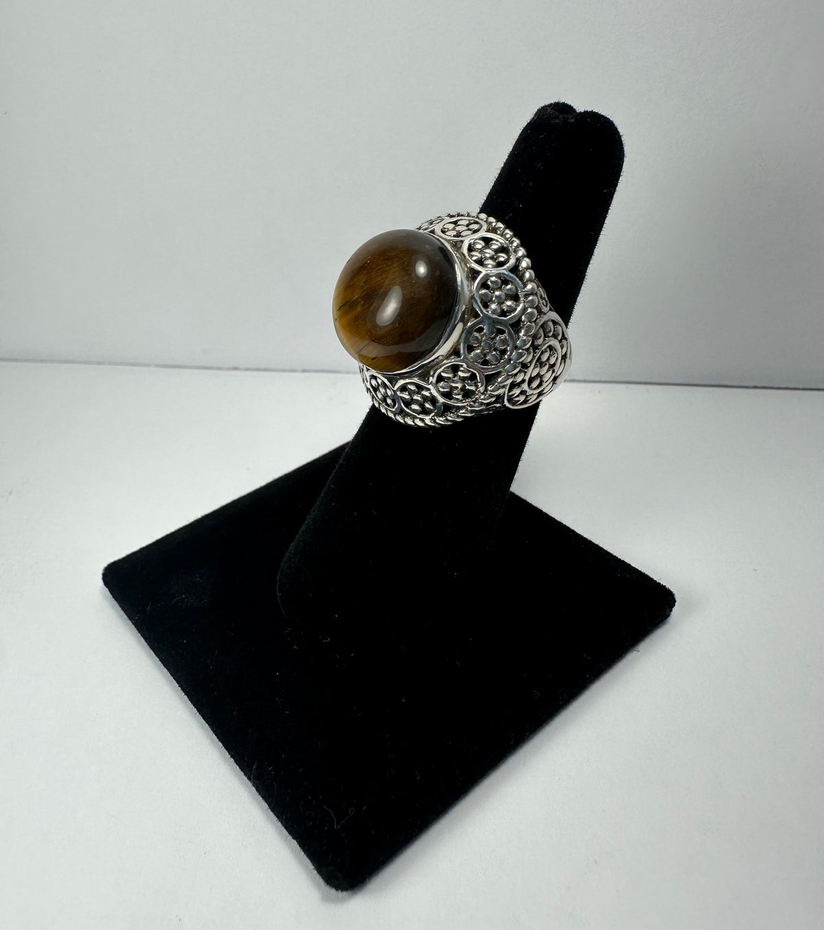 Silver ring with an ornate filigree pattern featuring circular and swirled details, topped with a polished round brown tiger's eye stone, displayed on a black ring stand.