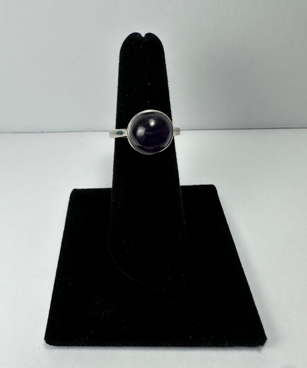 Silver ring with a round, polished deep purple amethyst stone set in a simple bezel, displayed on a black velvet ring holder against a white background.
