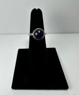 Silver ring with a round, polished deep purple amethyst stone set in a simple bezel, displayed on a black velvet ring holder against a white background.