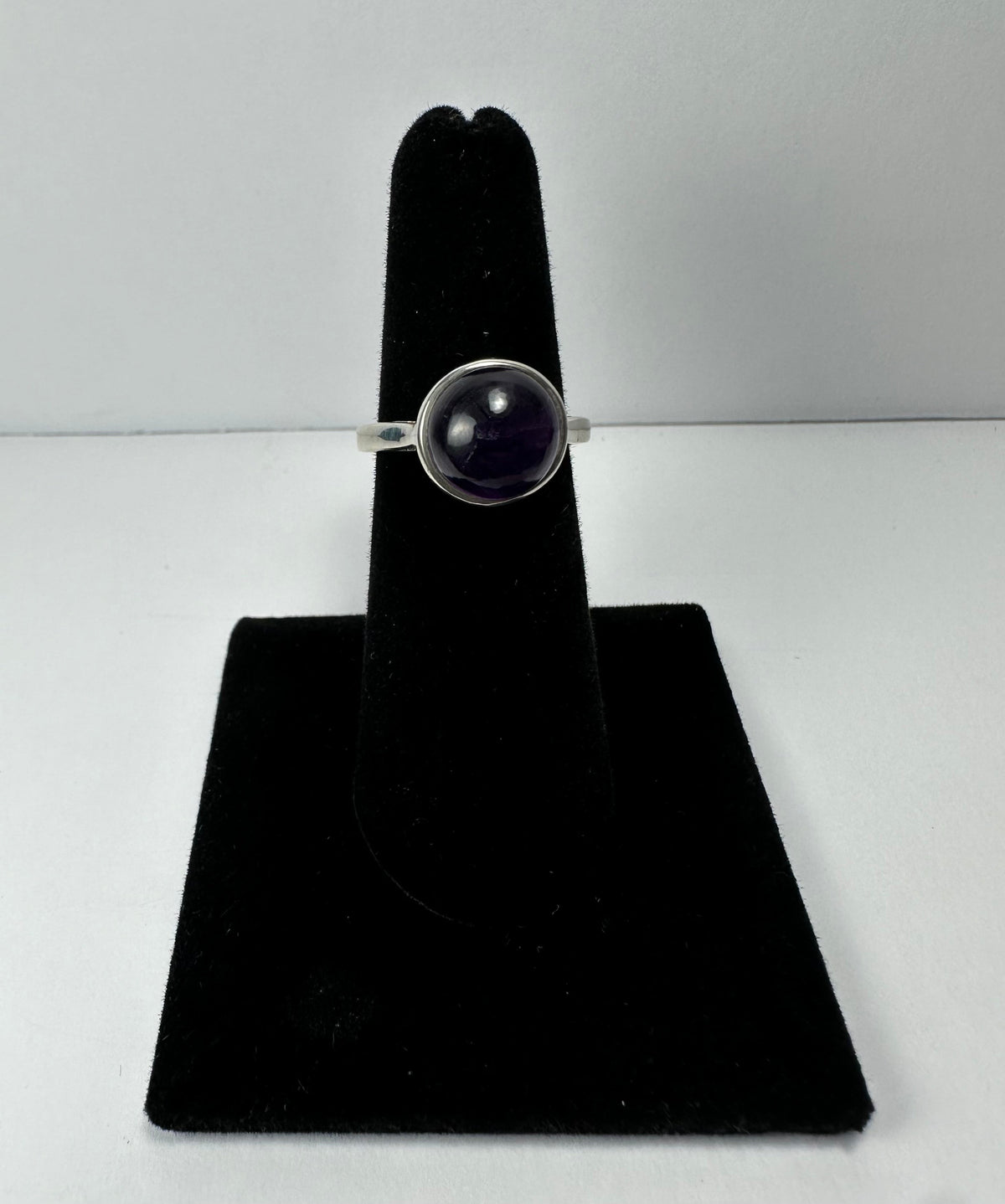 Silver ring with a round, polished deep purple amethyst stone set in a simple bezel, displayed on a black velvet ring holder against a white background.