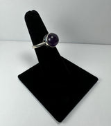 Silver ring with a round, polished deep purple amethyst stone set in a bezel setting displayed on a black velvet ring holder against a white background.