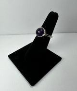 Silver ring with a round, polished deep purple amethyst stone set in a simple bezel on a black velvet display stand against a white background.