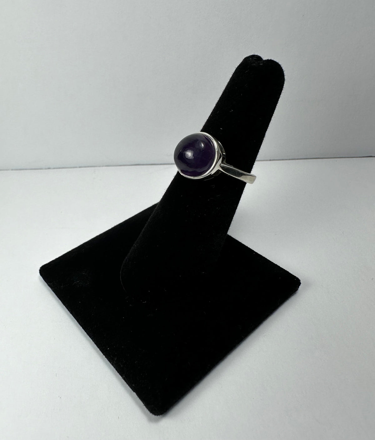 Silver ring with a round, polished deep purple amethyst stone set in a simple bezel on a black velvet display stand against a white background.